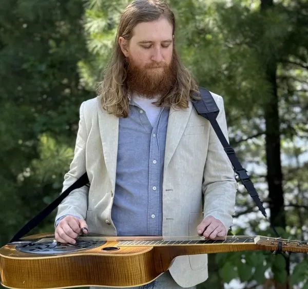 Adam Bachman playing a resonator guitar outdoors among pine trees